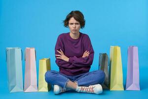 Severe young attractive short haired curly lady with natural makeup folding her hands on chest and frowning seriously eyebrows while looking at camera, isolated over blue background photo