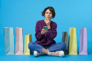 Thoughtful young brown-eyed curly brunette female holding smartphone and frowning eyebrows while looking pensively upwards, isolated over blue background with purchases photo