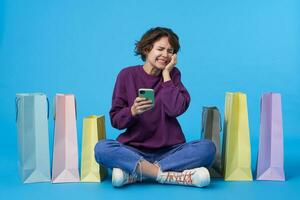 Good looking young curly brunette lady with casual hairstyle closing her eyes and frowning face while seeing something scaring on her mobile phone, isolated over blue background photo