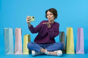 Indoor shot of lovely young curly brunette woman keeping her hand raised while making photo of herself and blowing air kiss at camera, posing over blue background