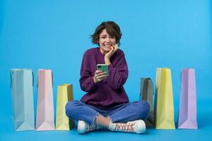 Studio photo of confused young curly brunette lady dressed in purple sweatshirt, jeans and sneakers showing her teeth while sitting over blue background with mobile phone