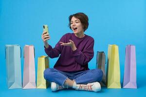 Positive young attractive curly brunette woman with short haircut looking joyfully at camera while making photo of herself on mobile phone, isolated over blue background