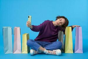 Pleased young lovely dark haired curly woman with natural makeup raising hand with mobile phone and smiling nicely at camera, sitting over blue background with paper bags photo