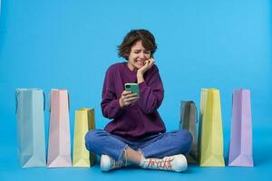 Afraid young attractive curly brunette woman keeping smartphone in raised hands and grimacing scaredly her face, sitting over blue background with paper bags photo