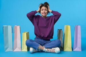 Afraid young brown-eyed short curly brunette woman clutching her head with raised hands and looking confusedly at camera, sitting over blue background with paper bags photo