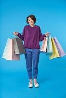 Pleased young lovely dark haired woman with short haircut raising hands with paper bags and looking positively at camera while shopping, isolated over blue background photo