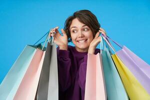 Positive young pretty dark haired curly woman in purple sweatshirt keeping shopping bags in raised hands and looking cheerfully aside, standing over blue background photo