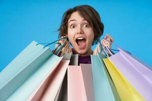 Bemused young pretty brown-eyed curly brunette lady raising her eyebrows while looking surprisedly at camera, raising her hands with purchases while posing over blue background photo