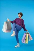 Energized young pretty short haired curly lady dancing happily while shopping and carrying many paper bags, isolated over blue background in casual clothes photo