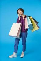 Confused young attractive short haired curly female grimacing her face while having phone conversation, standing over blue background with paper bags on her back photo