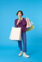 Puzzled young pretty short haired curly female keeping mobile phone in raised hand and looking pensively upwards, posing over blue background with shopping bags photo