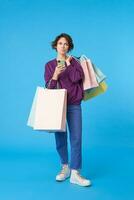 Pensive young pretty curly brunette woman squinting her eyes while looking thoughtfully aside and holding mobile phone, standing over blue background with shopping bags photo