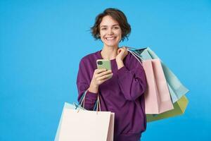 Portrait of happy young brown-eyed curly brunette lady smiling pleasantly at camera while standing over blue background with shopping bags and mobile phone in raised hands photo