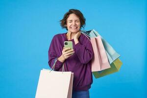 Pleasant looking young cheerful short haired curly female keeping her eyes closed while rejoicing about something, standing over blue background with shopping bags photo