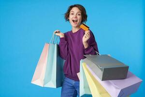 Portrait of joyful young brown-eyed curly brunette woman looking excitedly at camera while showing credit card, posing over blue background with shopping bags photo