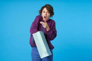 Surprised young brown-eyed dark haired woman with short haircut unpacking present and keeping her mouth opened, wearing purple sweatshirt and jeans while posing over blue background photo