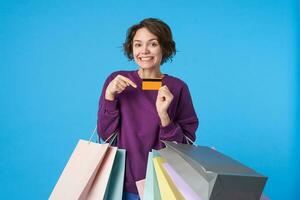 Portrait of young cheerful brown-eyed curly female with short haircut looking glady at camera while pointing on credit card in raised hand, standing over blue background with shopping bags photo