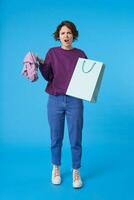 Disgruntled young lovely dark haired curly lady with natural makeup looking at camera with pout while keeping clothes and paper bag in raised hands, isolated over blue background photo