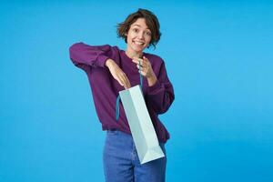 Indoor photo of young cheerful curly brunette female sticking hand inside of bag and looking excitedly at camera with surprised face, standing over blue background