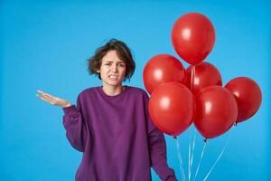 desconcertado joven de ojos marrones Rizado morena mujer con casual peinado ceñudo confusamente cara y levantamiento palma mientras en pie terminado azul antecedentes con aire globos foto