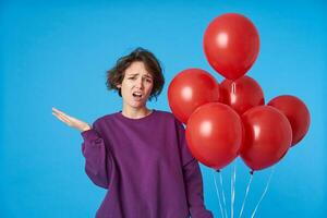 Puzzled young curly dark haired female raising confusedly hand and grimacing face while looking at camera, wearing purple sweatshirt while posing over blue background photo