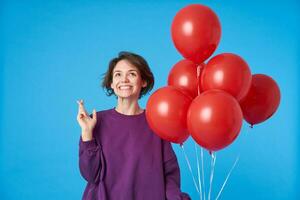 Happy young lovely dark haired lady smiling gladly and raising hand with crossed fingers while making wish on her birthday, standing over blue background with helium balloons photo