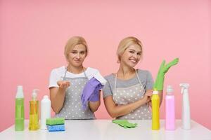 Studio photo of young blonde lovely housewives with natural makeup wearing rubber gloves while preparing for spring cleaning, posing over pink background
