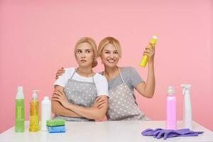 Indoor shot of young displeased pretty blonde female folding hands on chest while her happy lovely sister with detergent in raised hand embracing her, isolated over pink background photo