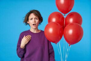 Confused young pretty short haired brunette woman frowning her eyebrows and holding palm on chest while looking at camera with puzzled face, posing over blue background photo