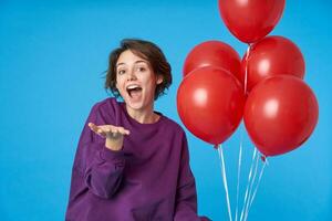 Overjoyed young lovely dark haired female with natural makeup looking excitedly at camera with broad smile and keeping palm raised, standing over blue background with air balloons photo