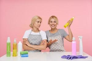 Happy young white-headed housewive in uniform raising bottle with detergent and looking cheerfully at camera while posing over pink background with her displeased colleague photo