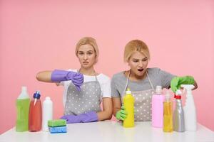 Displeased young blonde woman with natural makeup raising hand with dislike gesture while her excited white-headed amazed sister looking at bottles with household chemicals photo