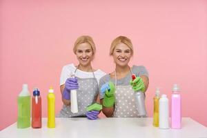 Cheerful young attractive white-headed cleaning ladies raising hands with spray bottles and looking happily at camera with broad smiles, sitting over pink background photo