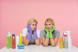 Indoor shot of young wailful blonde cleaning ladies holding their faces with raised hands and looking sadly on each other, having a lot of home work and making break photo