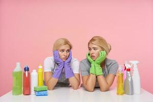 Sad young blonde cleaning ladies in rubber glove leaning their heands on raised hands and looking drearily on each other, sitting over pink background photo