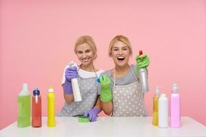 Happy young attractive blonde cleaning ladies dressed in uniform raising hands with spray bottles and smiling gladly at camera, isolated over pink background photo