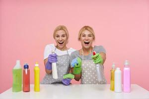 Overjoyed young blonde housewives in basic t-shirts and aprons keeping bottles with household chemicals and looking cheerfully at camera with excited faces, isolated over pink background photo