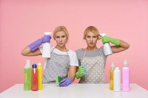 Puzzled young blonde ladies with casual hairstyle wearing rubber gloves and raising hands with atomizers to their temples, doing spring cleaning and making small break, isolated over pink background photo