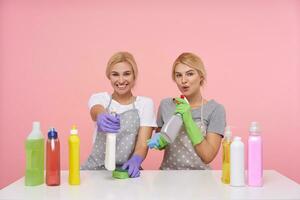 Indoor photo of lovely young white-headed housewives being in nice mood while making spring cleaning, sitting over pink background with household chemicals