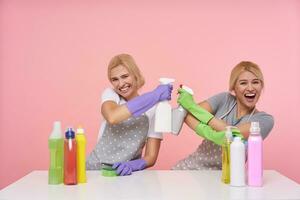 alegre joven rubia mujer con casual peinado sonriente extensamente mientras haciendo divertido, tomando descanso con limpieza casa y sentado terminado rosado antecedentes con botellas de detergentes foto