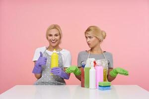 Indoor shot of young white-headed cleaning ladies sitting over pink background in working clothes, one getting best detergent and another has a lot of worse bottles photo