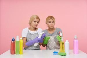 Bewildered young pretty blonde women dressed in uniform holding spray bottles with household chemicals and looking at camera with puzzled faces, isolated over pink background photo