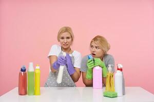 Confused young pretty blonde sisters dressed in working clothes making cleaning and posing over pink background with puzzled faces. Housework and housekeeping concept photo