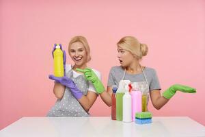 Bewildered young white-headed lady in green rubber gloves raising confusedly palms while looking at her cheerful blonde sister with yellow bottle in raised hand, isolated over pink background photo