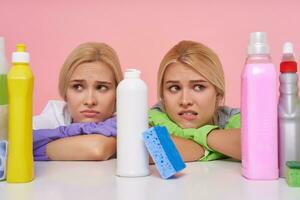 Portrait of unhappy young white-headed ladies keeping their heads on folded hands and looking tiredly on detergents bottles, sitting over pink background photo