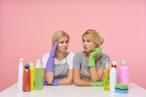 Weary young white-headed ladies dressed in uniform being tired after spring cleaning and looking sadly on each other, sitting over pink background with household chemicals photo