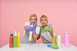 Afraid young white-headed cleaning ladies defending theirselves with spray bottles of detergents, seeing thieves while making cleaning, posing over pink background photo