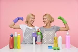 Glad young attractive blonde housewives rairing hands in multi-colored rubber glovves and looking happily on ech other with broad smile, sitting over pink background with detergents photo