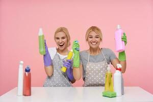 Cheerful lovely young blonde women wearing uniform while cleaning house, sitting over pink background with detergents and smiling joyfully at camera, being happy to finish their work photo