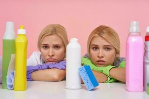 Portrait of young sad white-headed ladies wearing multi-colored rubber gloves while making cleaning, looking sorrowfully at camera and keeping heads on folded hands, isolated over pink background photo